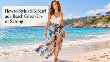 Woman styling an Aubergine XL silk scarf as a beach cover-up and sarong beside turquoise water.