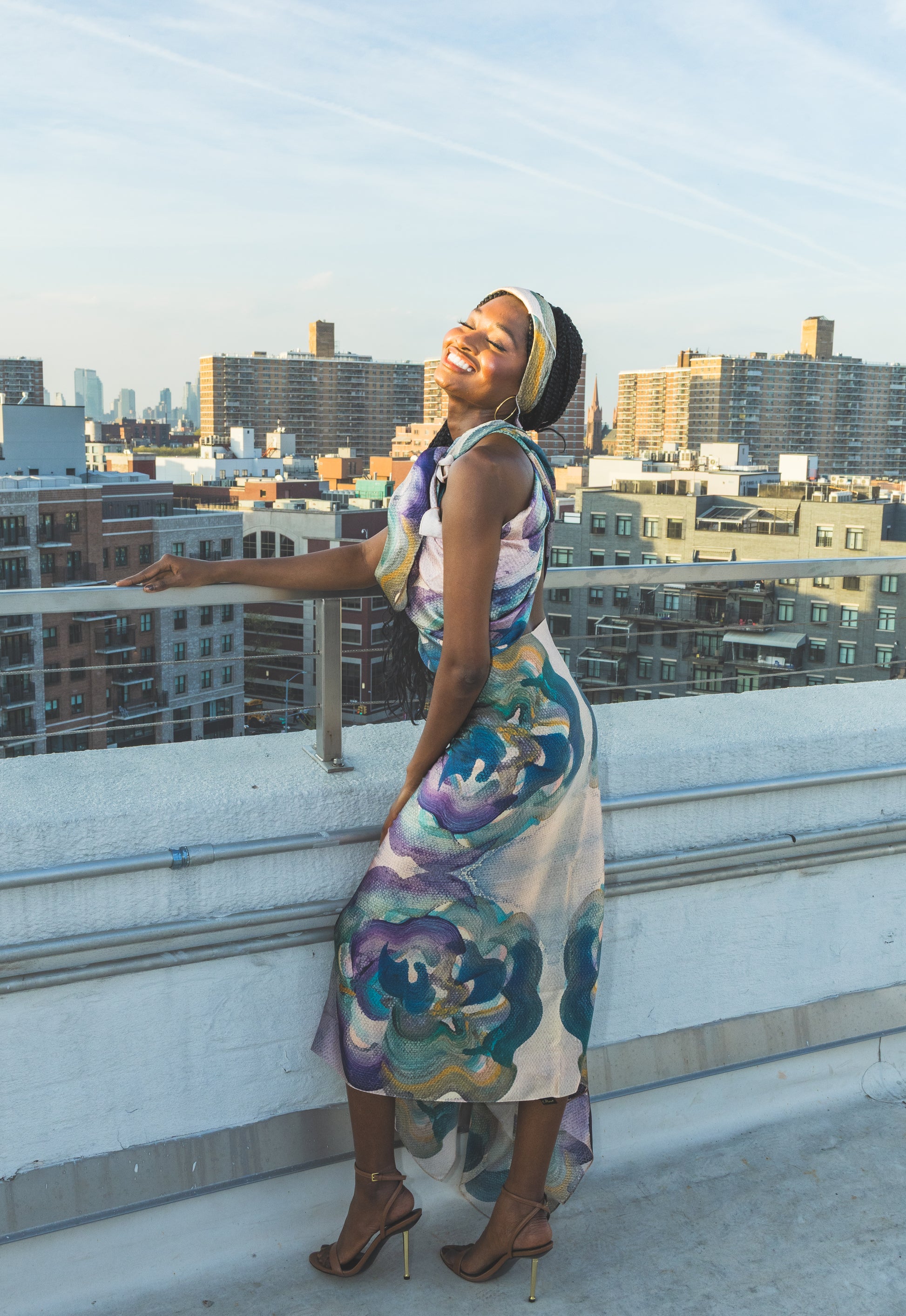 A model poses on a rooftop in a flowing floral dress with soft blue, green, and purple tones. The city skyline and warm sunlight create a bright, stylish, and elegant fashion look.