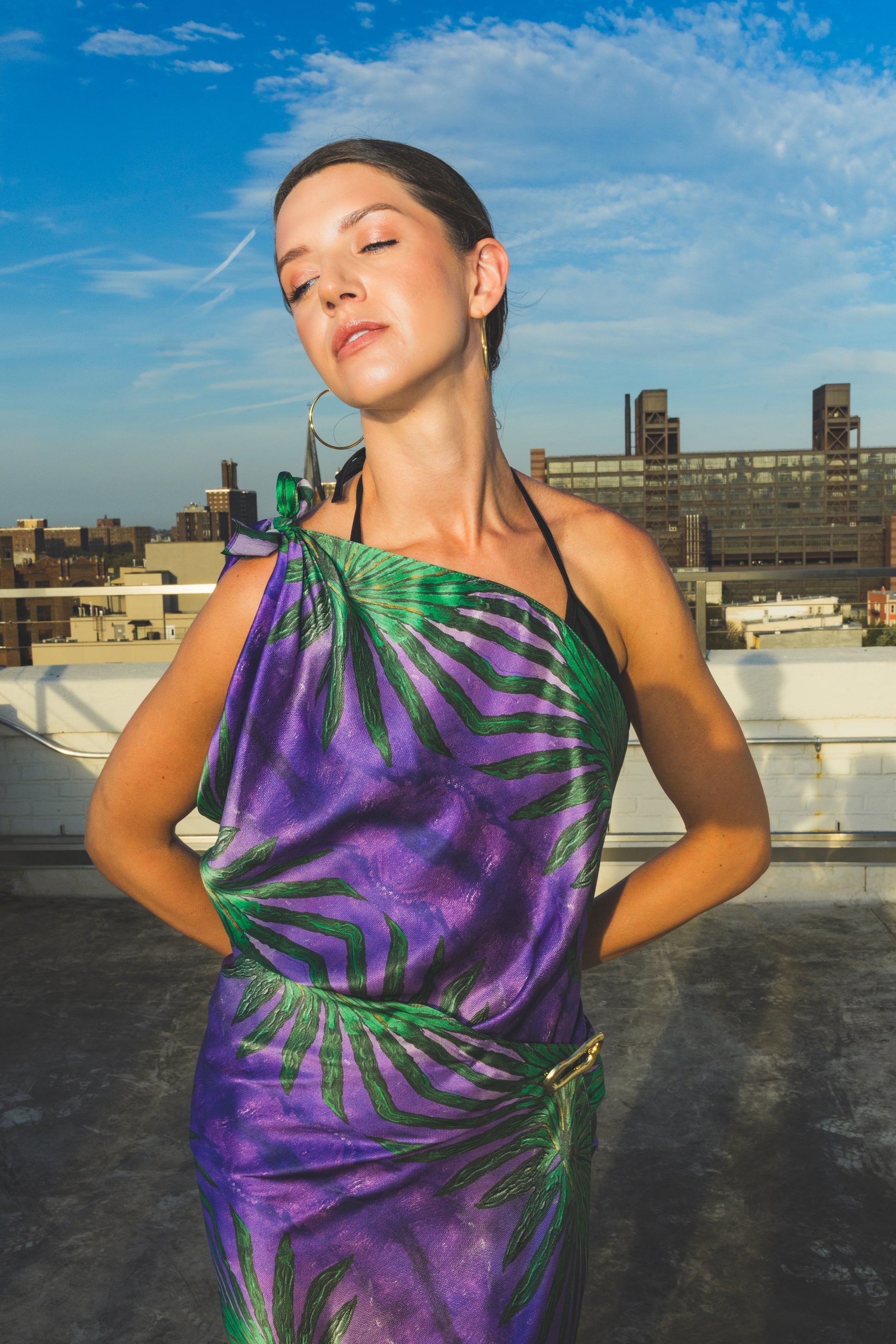 A model poses on a rooftop wearing a vibrant purple and green one-shoulder dress with a bold tropical print, creating a stylish and confident editorial fashion look.