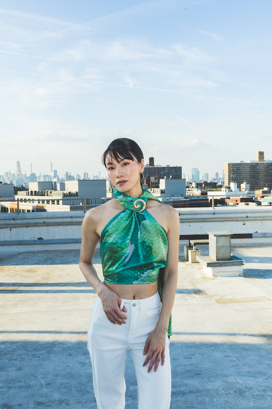 A model poses on a rooftop wearing a shiny green halter scarf top with white pants, creating a fresh, elegant, and modern city-fashion look.