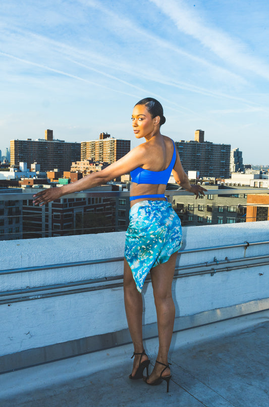 A model sits on an industrial rooftop wearing green abstract-print pants with a matching headscarf and white tank top, creating a fresh, relaxed urban fashion look.