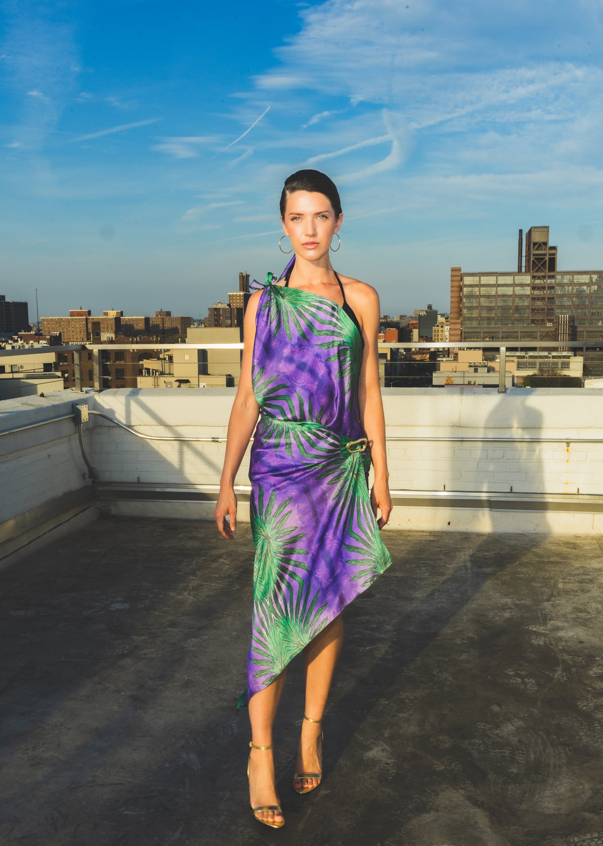 A model walks confidently on a rooftop wearing a purple one-shoulder dress with bold green tropical leaf patterns, creating a vibrant and elegant fashion look.