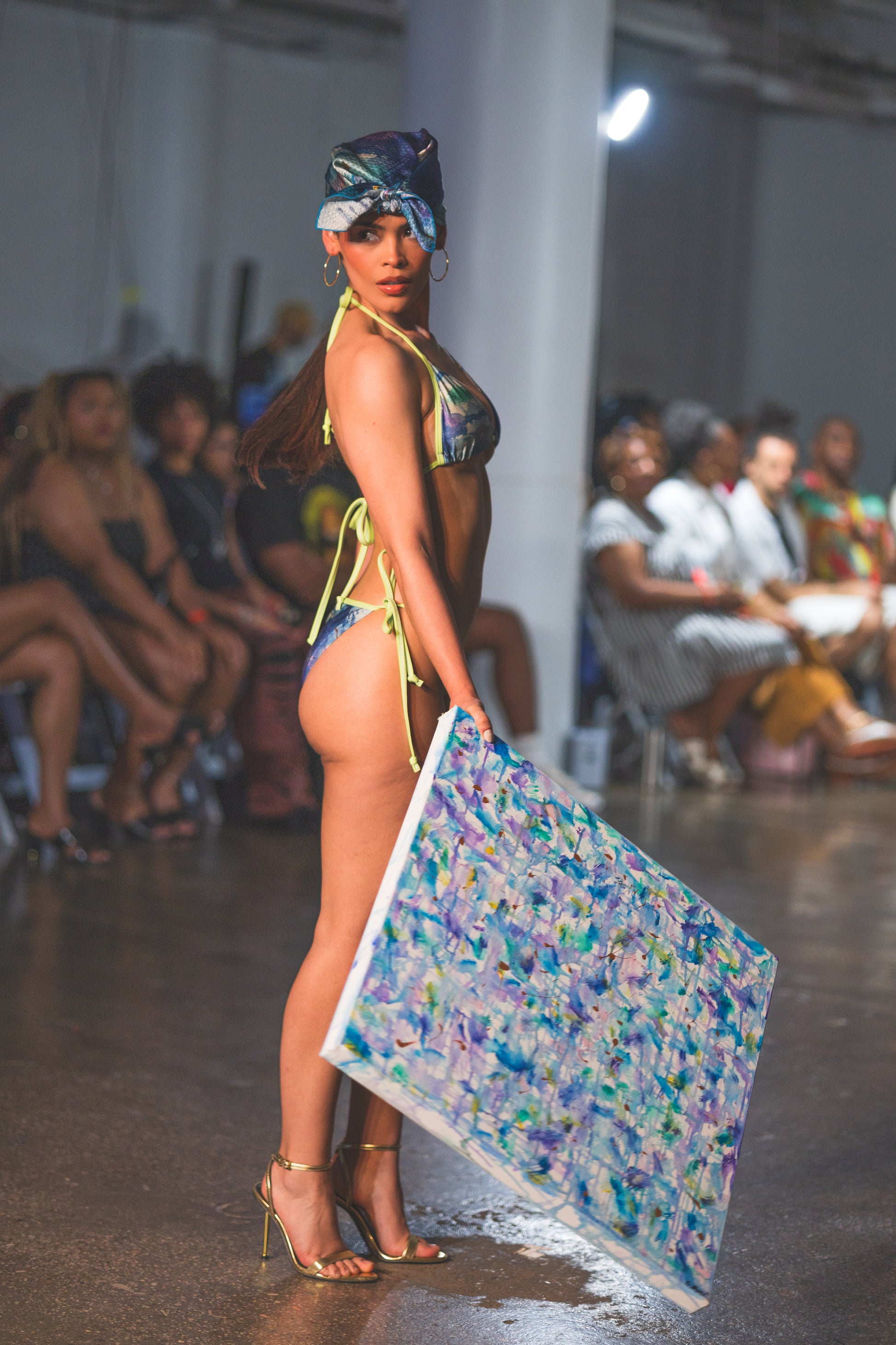 A runway model poses in a colorful bikini with a matching headwrap, holding a large abstract artwork. The look feels bold, artistic, and high-fashion.