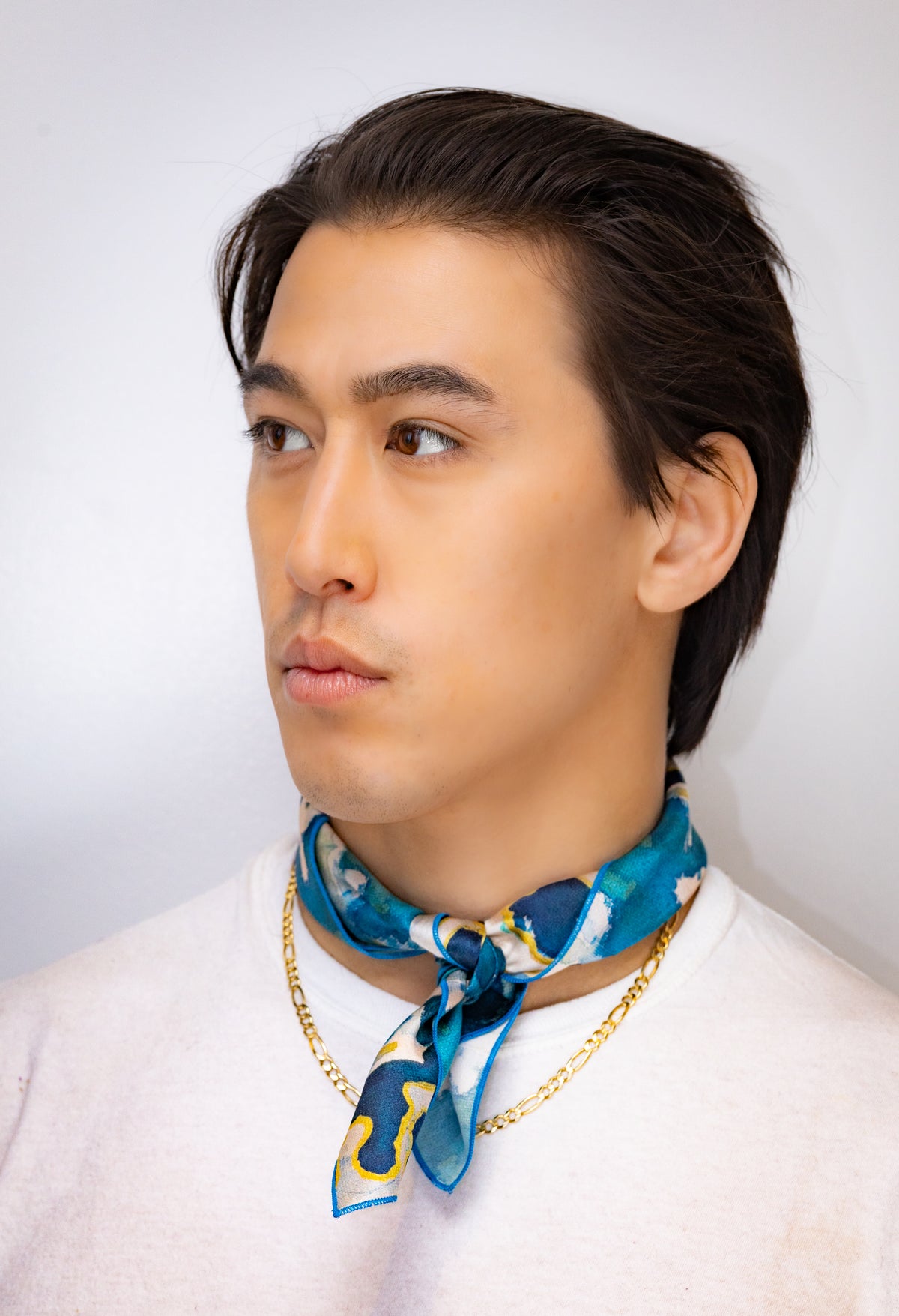 Man wearing a blue patterned scarf and gold chain necklace against a white background