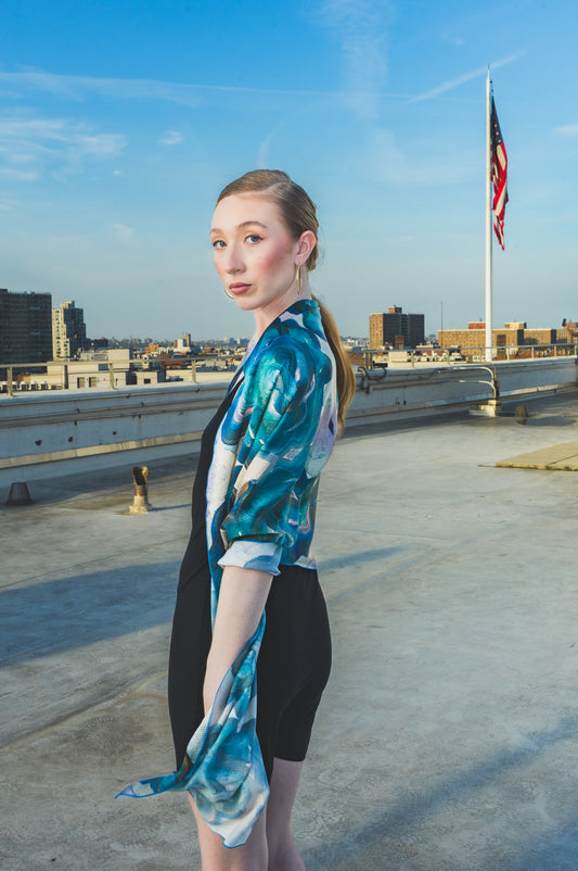 A model poses on a rooftop wearing a black dress styled with a sheer blue-green floral scarf wrap, creating an elegant and artistic city-fashion look.