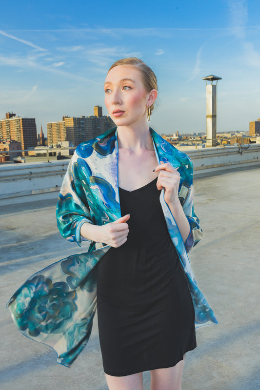 A model poses on a rooftop wearing a black dress styled with a sheer blue-green floral scarf wrap, creating an elegant and artistic city-fashion look.