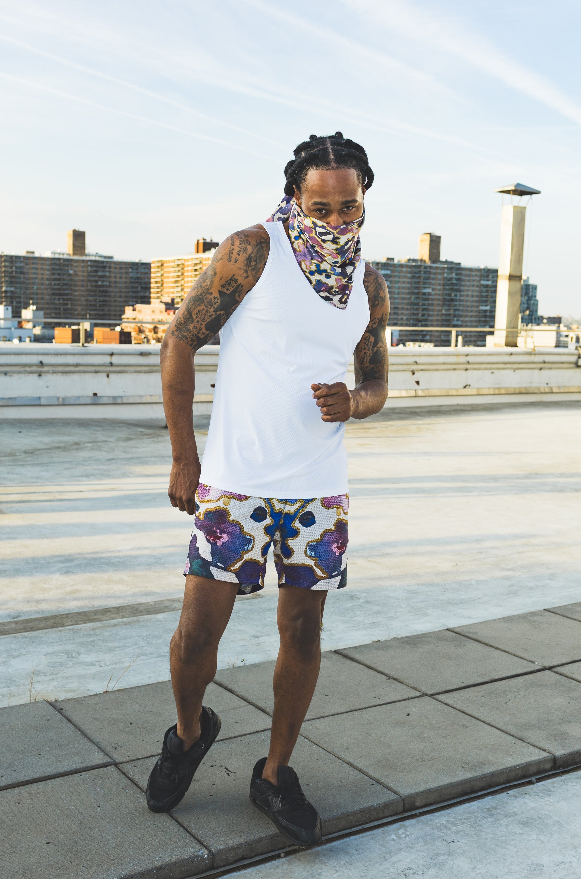 A male model poses on a rooftop wearing a white tank top, abstract-print shorts, and a matching face scarf, creating a bold urban streetwear fashion look.