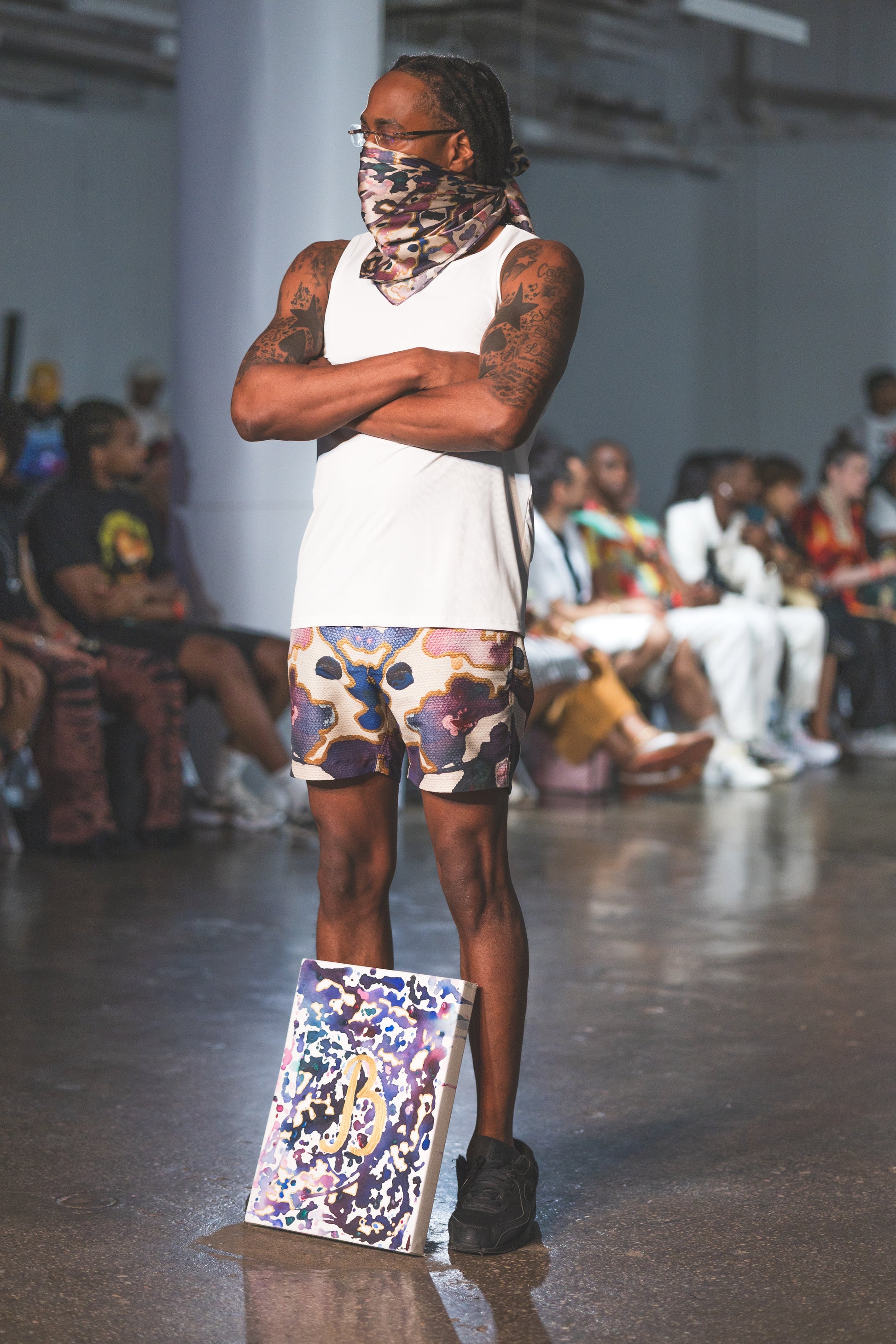 A male runway model stands confidently in a white tank top, abstract-print shorts, and matching face scarf, paired with black shoes for a bold urban fashion look.