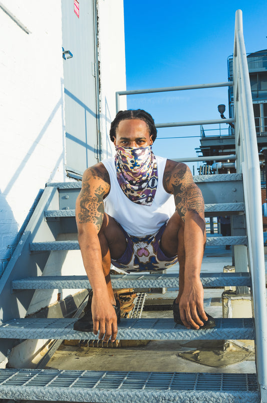 A male model poses on outdoor metal stairs wearing a white tank top, abstract-print shorts, and a matching face scarf, creating a bold urban streetwear look.
