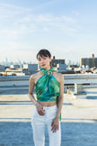 A model poses on a rooftop wearing a shiny green halter scarf top with white pants, creating a fresh, elegant, and modern city-fashion look.