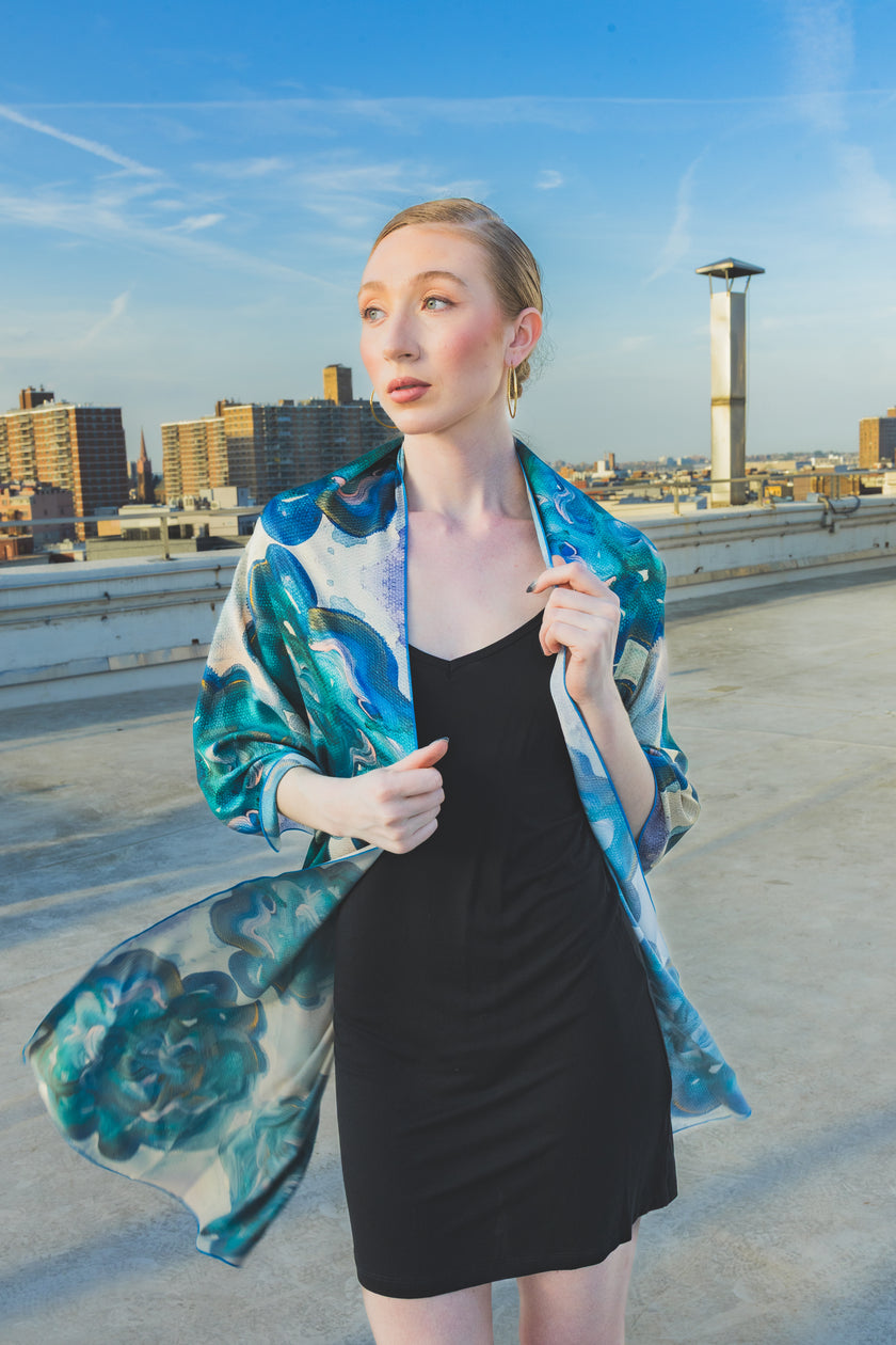 A model poses on a rooftop wearing a black dress styled with a sheer blue-green floral scarf wrap, creating an elegant and artistic city-fashion look.