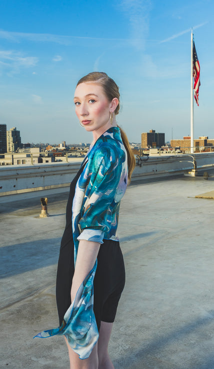A model poses on a rooftop wearing a black dress styled with a sheer blue-green floral scarf wrap, creating an elegant and artistic city-fashion look.
