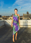 A model walks confidently on a rooftop wearing a purple one-shoulder dress with bold green tropical leaf patterns, creating a vibrant and elegant fashion look.