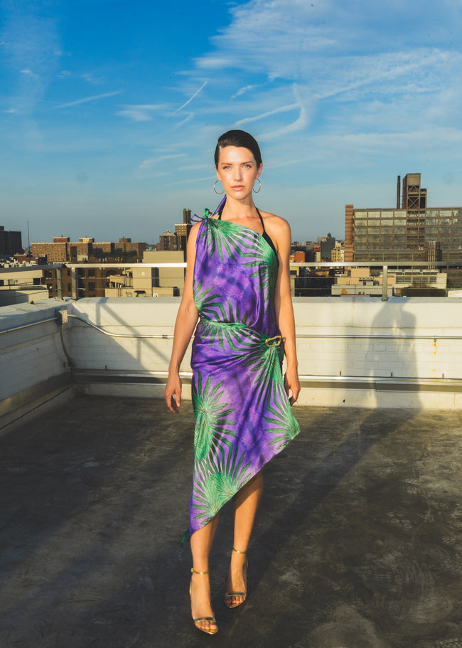 A model walks confidently on a rooftop wearing a purple one-shoulder dress with bold green tropical leaf patterns, creating a vibrant and elegant fashion look.