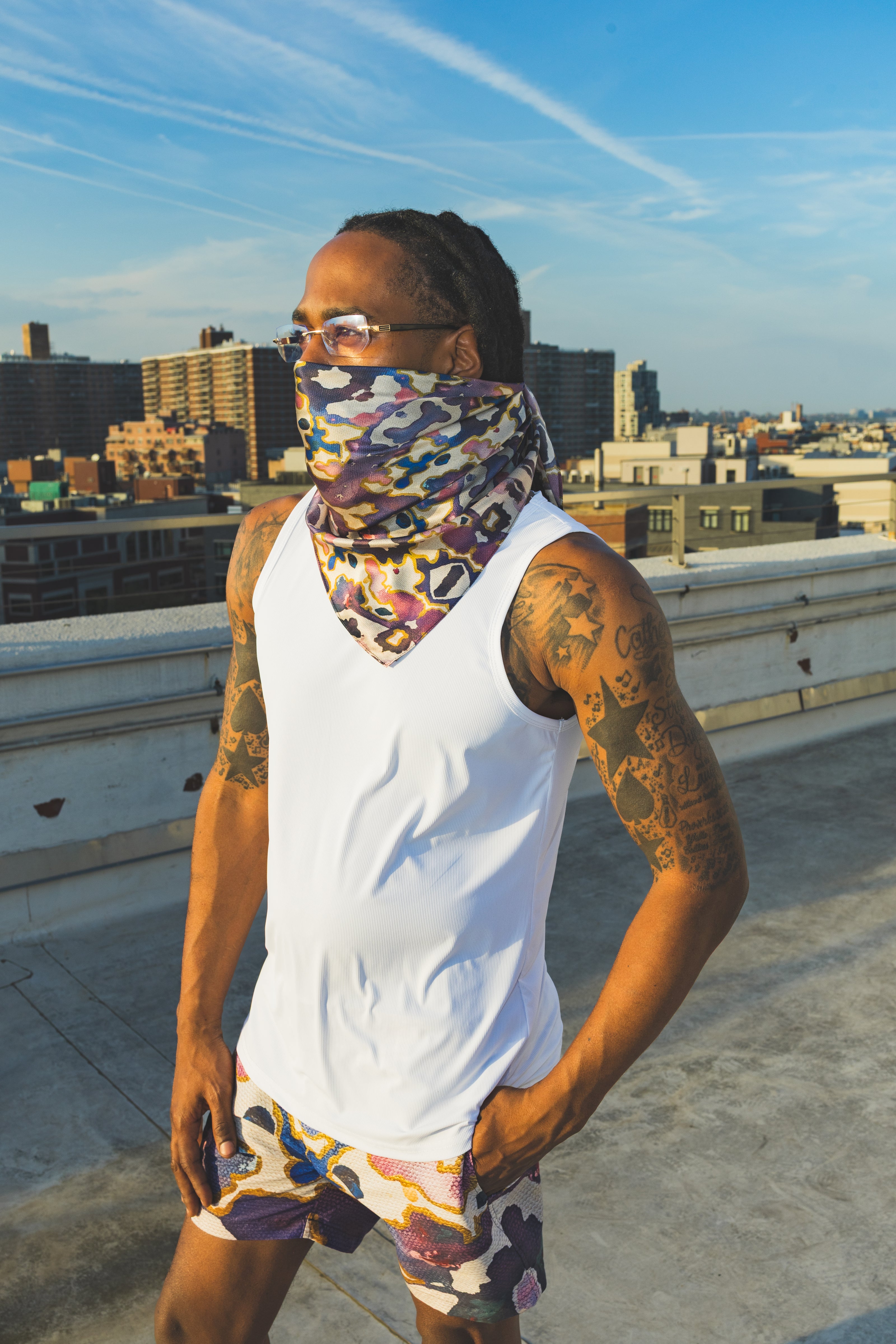 A male model poses on a rooftop wearing a white tank top with an abstract-print face scarf and matching shorts, creating a bold urban streetwear look.