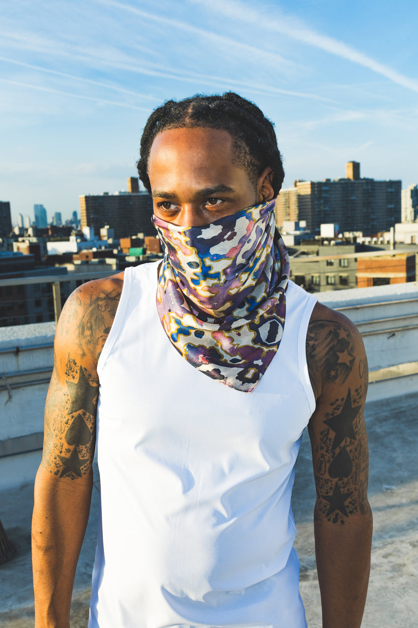 A male model poses on a rooftop wearing a white tank top and an abstract-print scarf mask, creating a bold streetwear-inspired fashion look with an urban city backdrop.