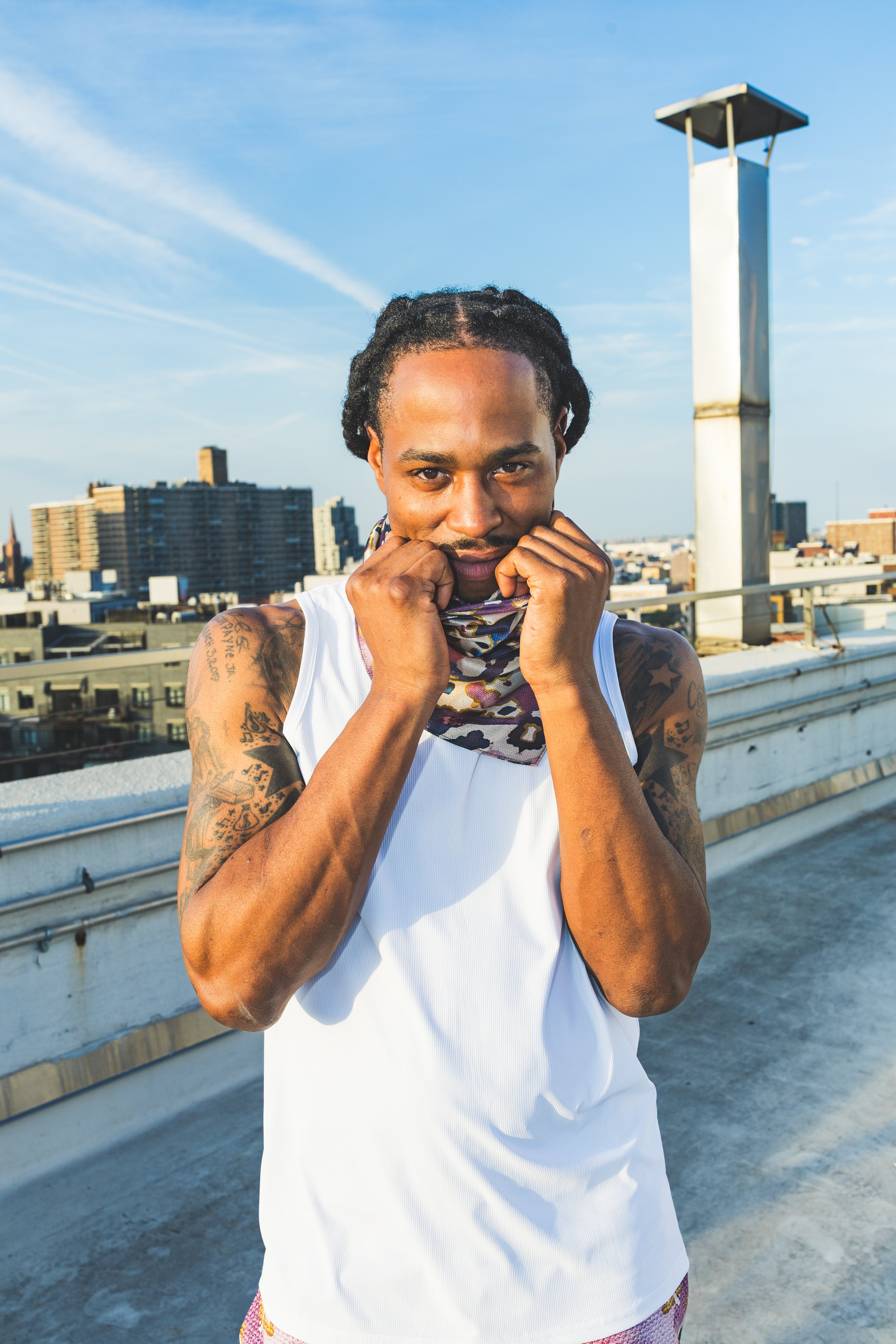 A male model poses on a rooftop wearing a white tank top with abstract-print shorts, creating a casual, confident, and stylish streetwear look.