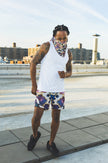 A male model poses on a rooftop wearing a white tank top, abstract-print shorts, and a matching face scarf, creating a bold urban streetwear fashion look.
