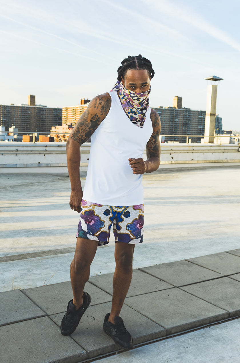 A male model poses on a rooftop wearing a white tank top, abstract-print shorts, and a matching face scarf, creating a bold urban streetwear fashion look.