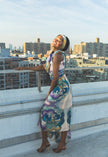 A model poses on a rooftop in a flowing floral dress with soft blue, green, and purple tones. The city skyline and warm sunlight create a bright, stylish, and elegant fashion look.