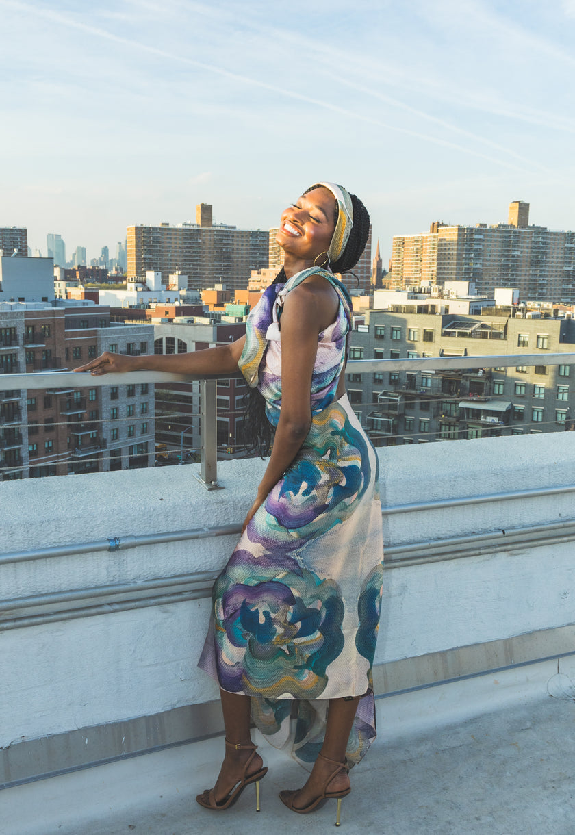 A model poses on a rooftop in a flowing floral dress with soft blue, green, and purple tones. The city skyline and warm sunlight create a bright, stylish, and elegant fashion look.