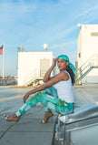 A model sits on an industrial rooftop wearing green abstract-print pants with a matching headscarf and white tank top, creating a fresh, relaxed urban fashion look.
