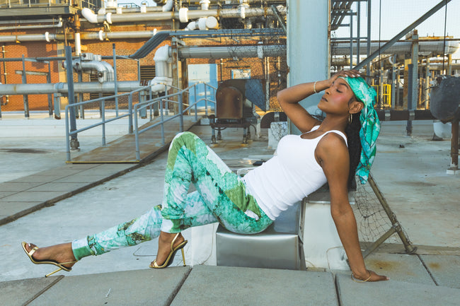 A model poses in green abstract-print pants with a matching headscarf and white tank top, creating a relaxed urban fashion look against an industrial rooftop setting.