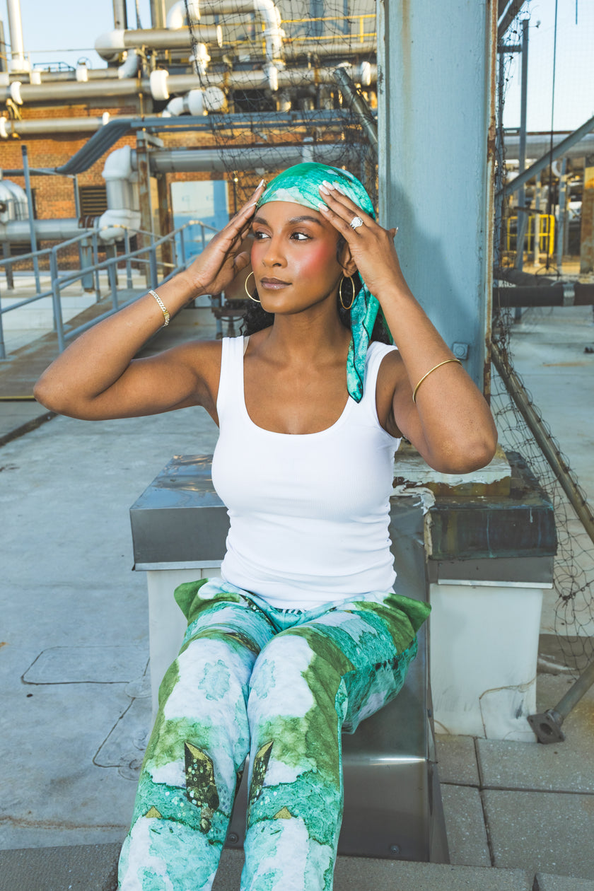 A model sits in a white tank top with green printed pants and a matching headscarf, creating a fresh, stylish, and relaxed urban fashion look.