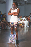A male runway model stands confidently in a white tank top, abstract-print shorts, and matching face scarf, paired with black shoes for a bold urban fashion look.