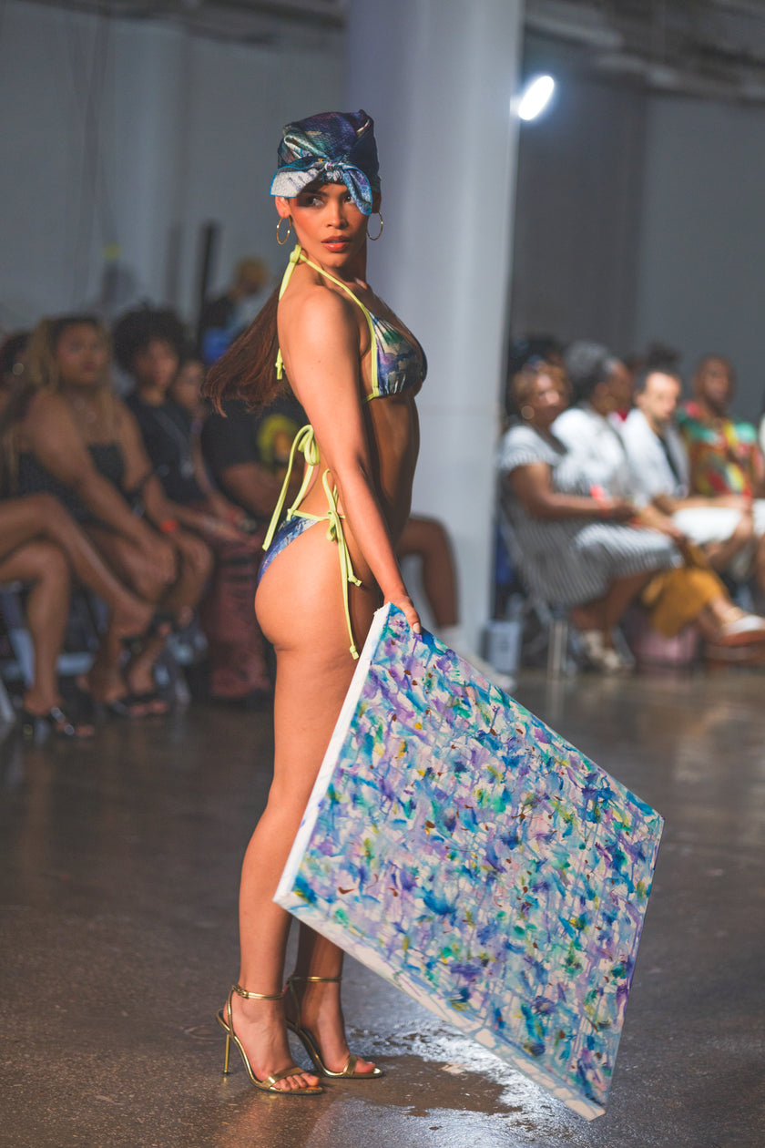 A runway model poses in a colorful bikini with a matching headwrap, holding a large abstract artwork. The look feels bold, artistic, and high-fashion.