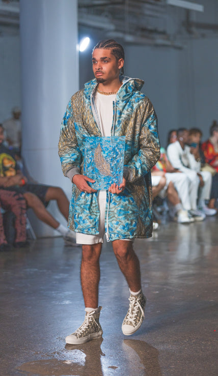Man walking on a runway wearing a blue and gray patterned jacket at a fashion show.