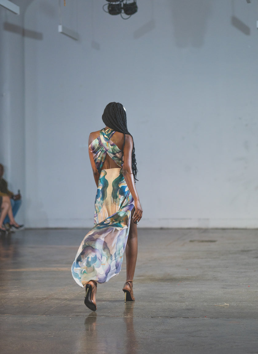 A runway model showcases a flowing floral maxi dress with an open-back design and high slit. The soft watercolor-style print and graceful movement give the image a refined, elegant fashion-show feel.