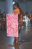 A runway model walks in a strapless floral dress while carrying a large pink abstract artwork. The look feels bold, artistic, and high-fashion with an editorial runway mood.