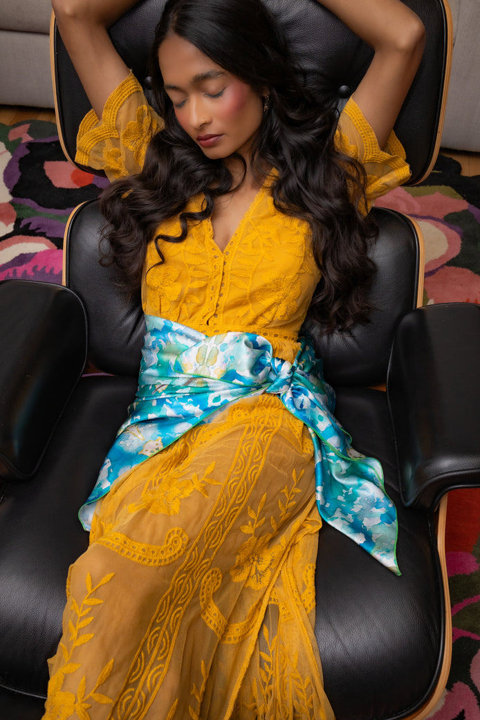 Woman in a yellow dress with a blue scarf sitting on a black chair.
