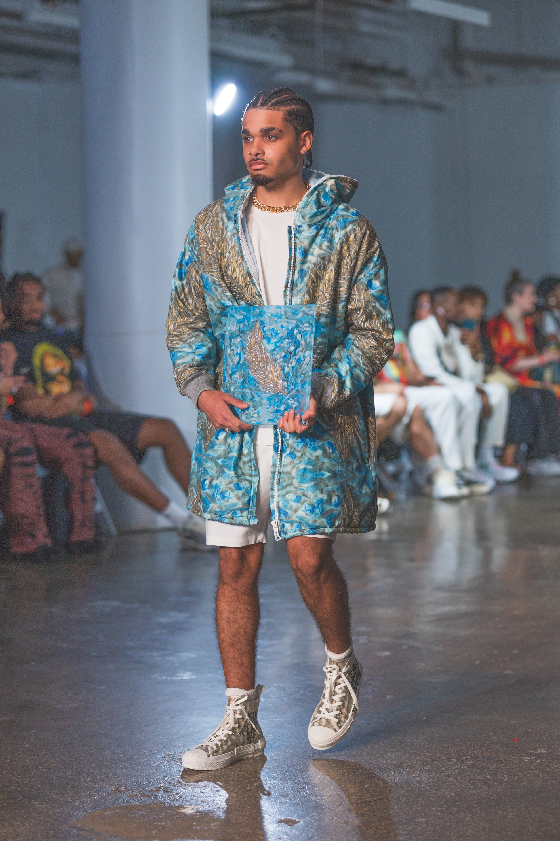 Man walking on a runway wearing a blue and gray patterned jacket at a fashion show.