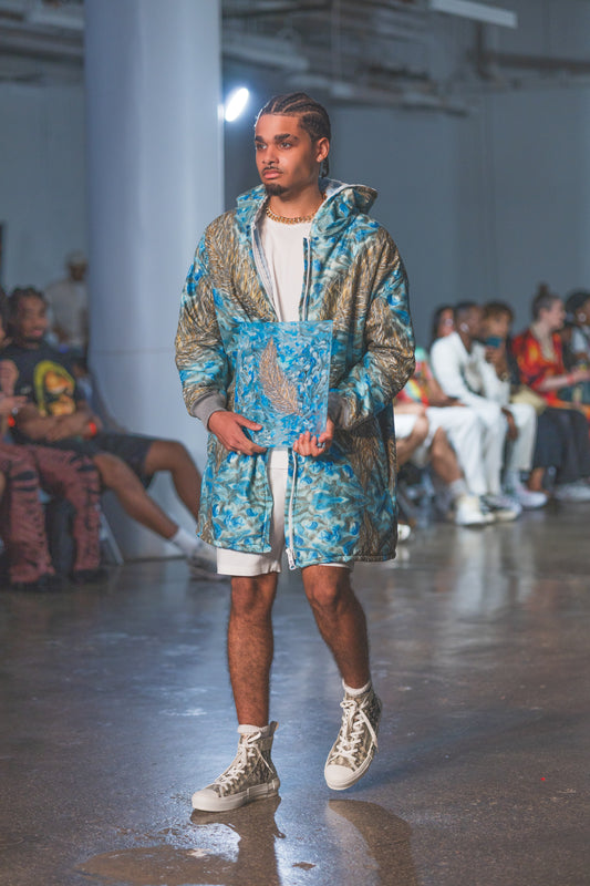Man walking on a runway wearing a blue and gray patterned jacket at a fashion show.