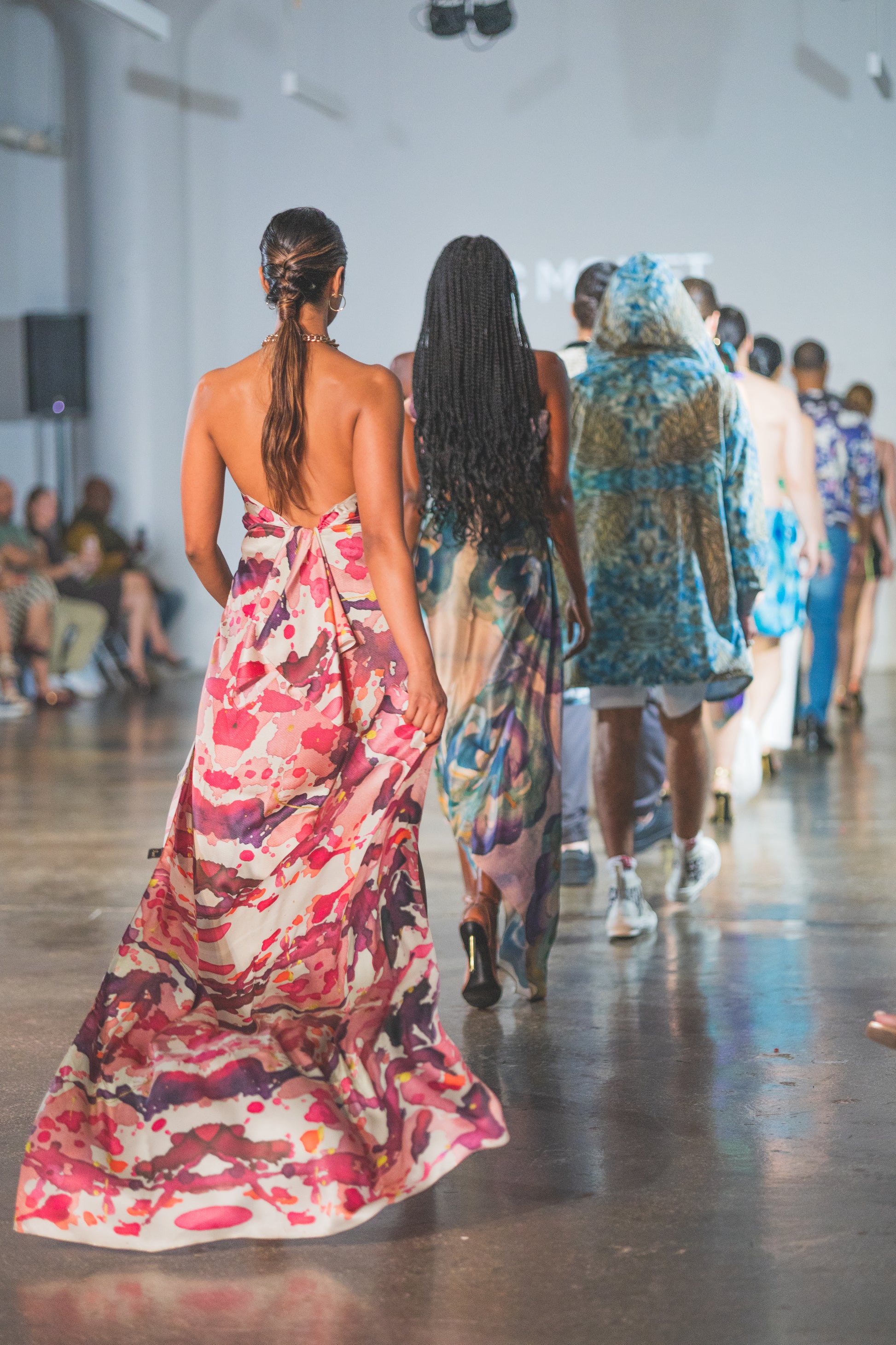 Model wearing a colorful floral dress walking on a fashion show runway.
