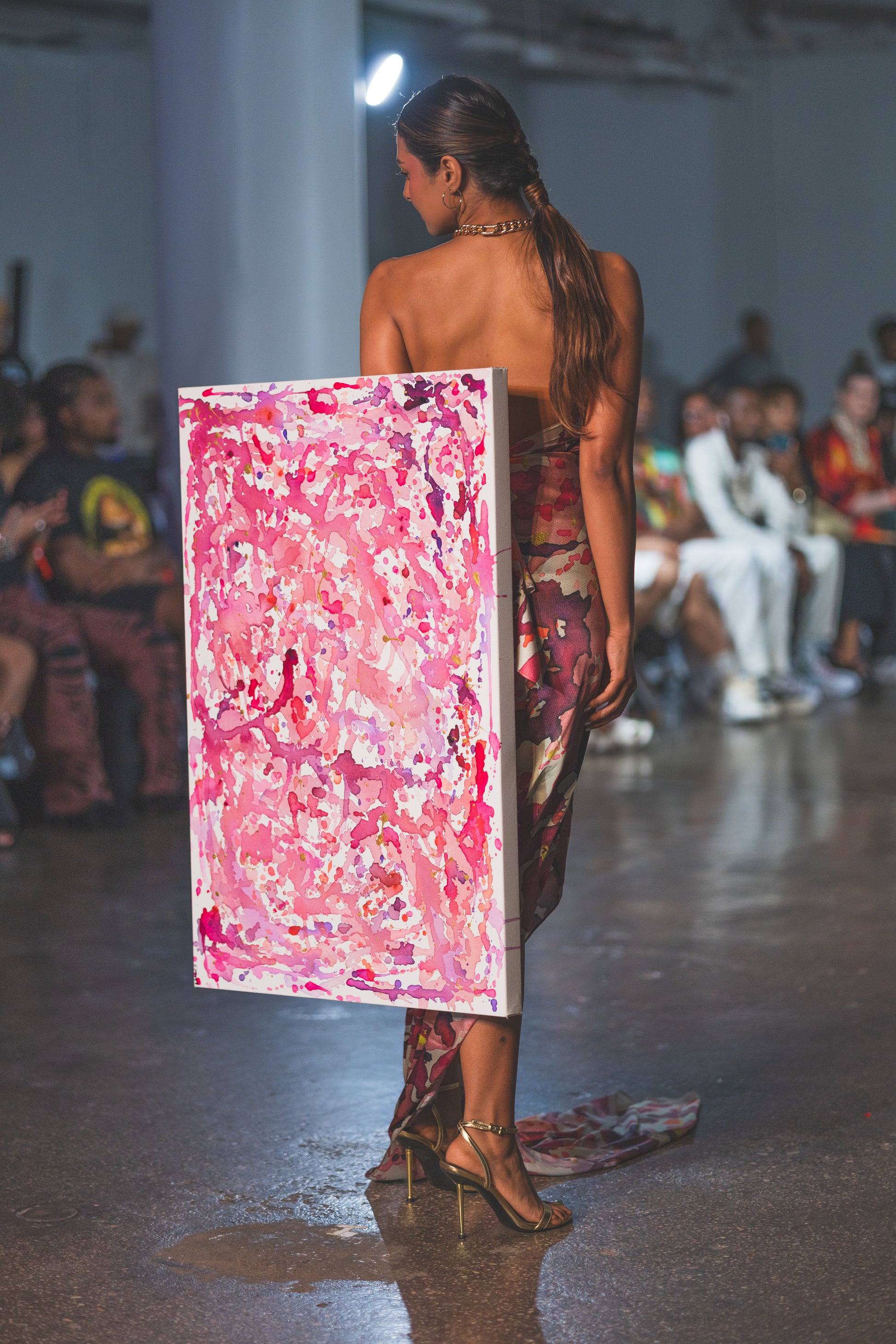 A runway model walks in a strapless floral dress while carrying a large pink abstract artwork. The look feels bold, artistic, and high-fashion with an editorial runway mood.