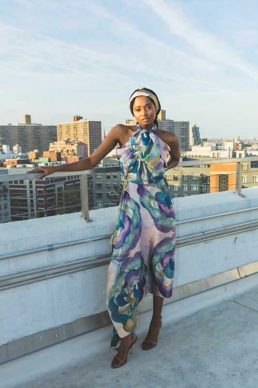 A model poses on a rooftop in a vibrant floral halter dress with soft purple, blue, and green tones. The city skyline and golden sunlight create a chic, modern fashion look.
