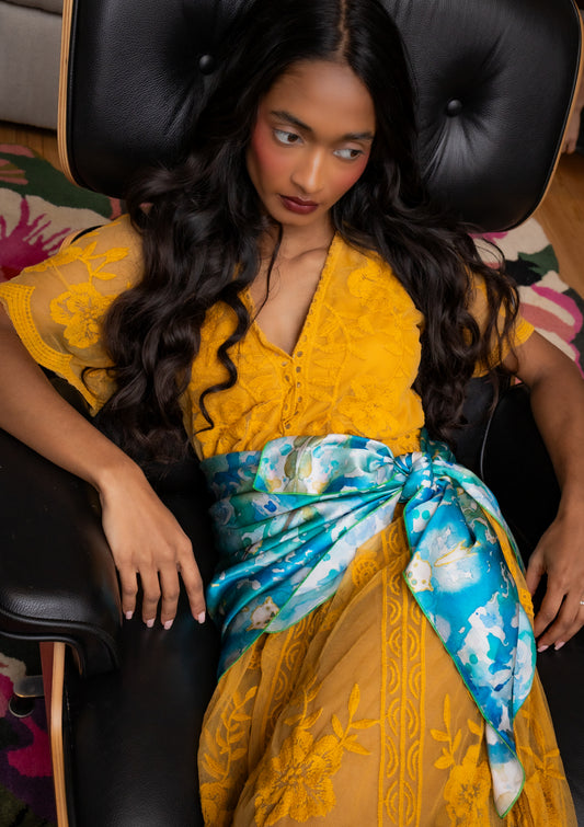 Woman in a yellow dress with a blue floral scarf sitting on a black chair.