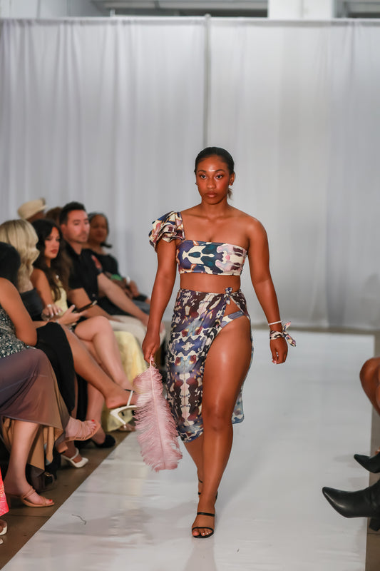 Model wearing the Andromeda one shoulder bikini top with printed bottoms and a wrap on a runway.