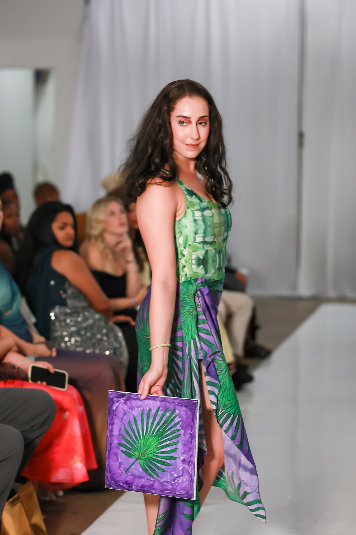 Runway model in a women's green floral one piece swimsuit styled with a purple wrap skirt, carrying a purple palm-print clutch