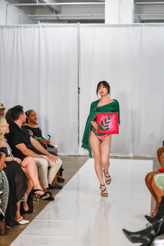 Runway model walking in a pink floral one piece swimsuit with a green silk wrap over her shoulders, carrying a pink leaf-print clutch