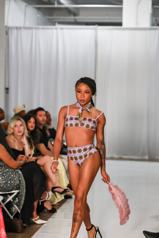 Runway model walking in a women's bralette bikini top with matching high-waist bottoms in a lavender and brown circle print, holding a pink feather fan