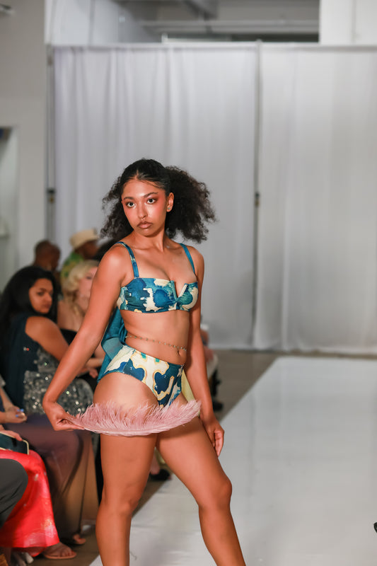 Runway model in a C. Monet Atelier blue and cream floral-print bralette bikini set, holding a pink feather fan at her hips on a white runway