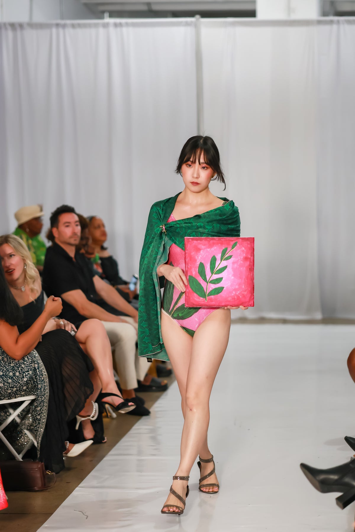 Front view of a designer one piece swimsuit in pink with a green leaf print, styled with a green wrap on the C. Monet Atelier runway