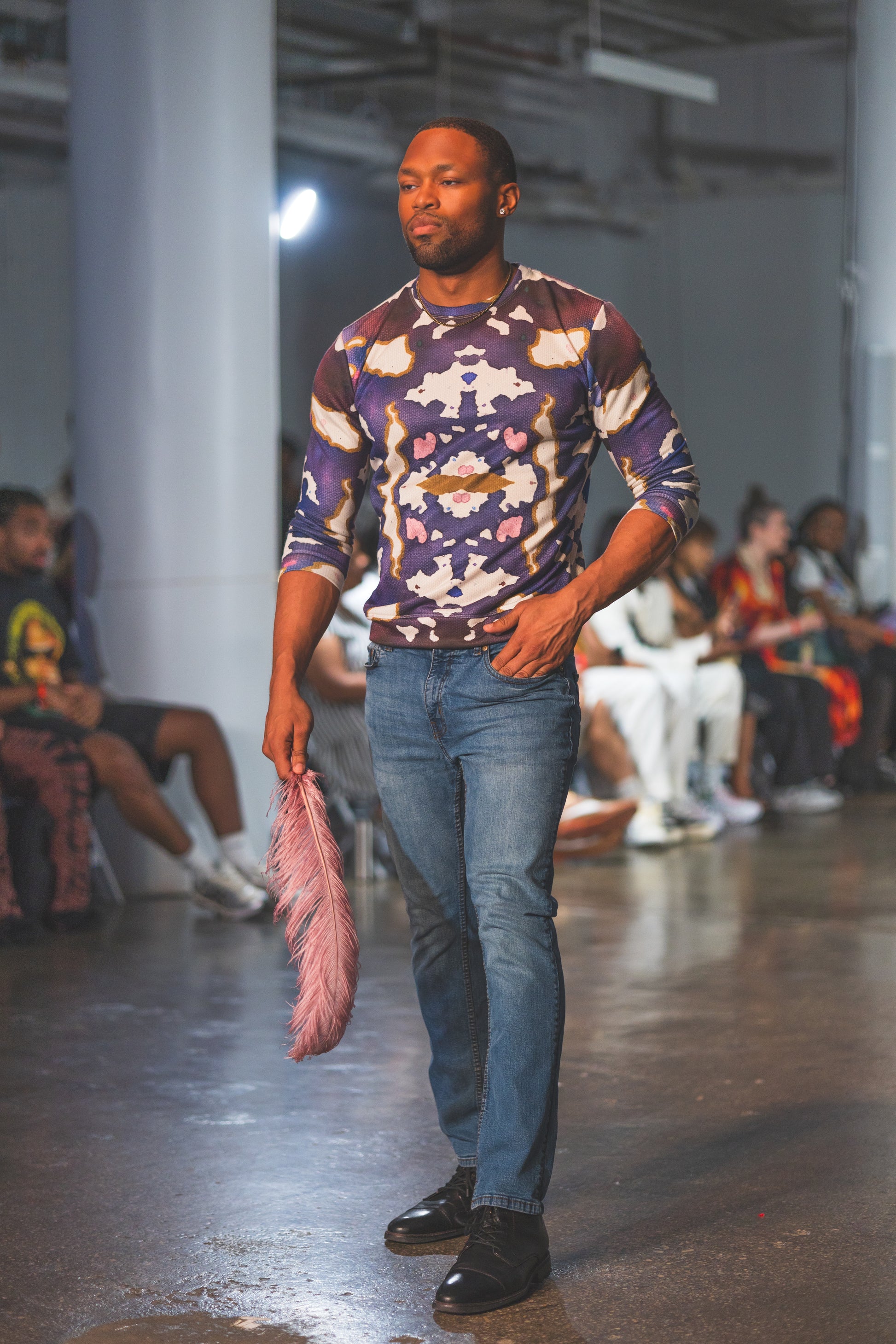 A male runway model walks confidently wearing a bold abstract-print long-sleeve top paired with blue jeans and black shoes, creating a stylish modern fashion look.