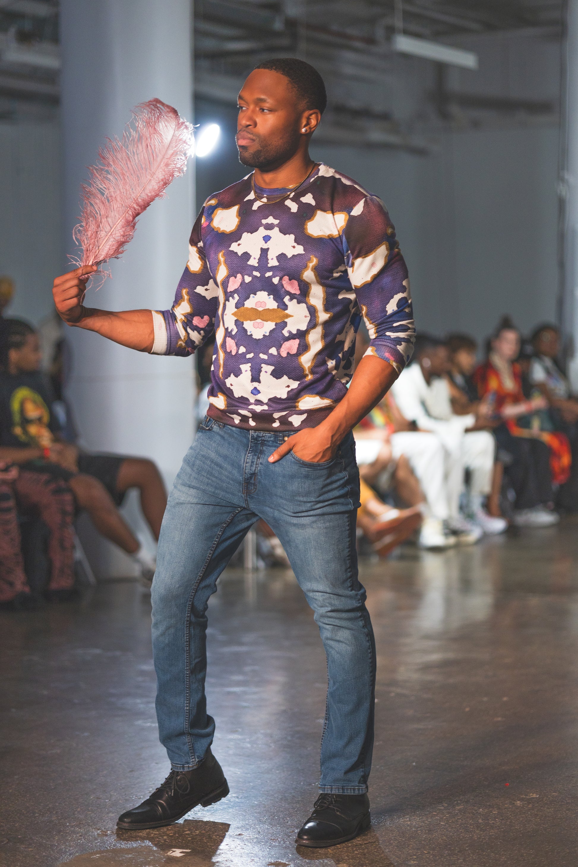 Man on a fashion runway wearing a patterned shirt and jeans, holding pink smoke.
