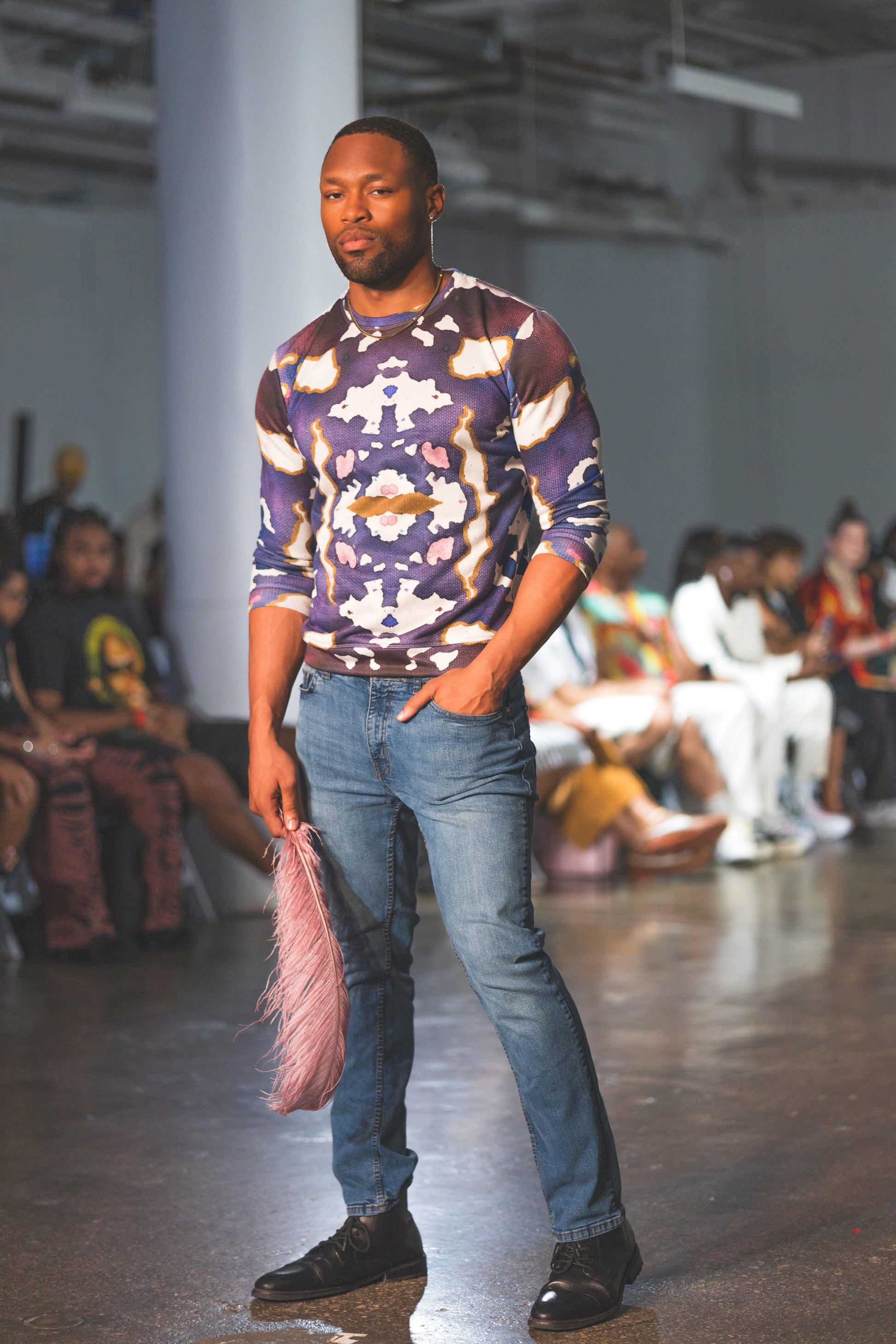 A male runway model walks confidently wearing a bold abstract-print long-sleeve top paired with blue jeans and black shoes, creating a stylish modern fashion look.
