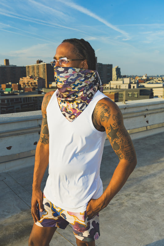A male model poses on a rooftop wearing a white tank top with an abstract-print face scarf and matching shorts, creating a bold urban streetwear look.